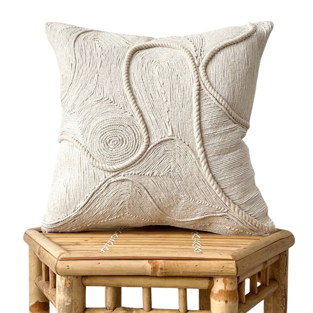 Studio shot of the Arshi cushion with abstract, organic cord appliqué design in ivory, displayed on a woven rattan stool.