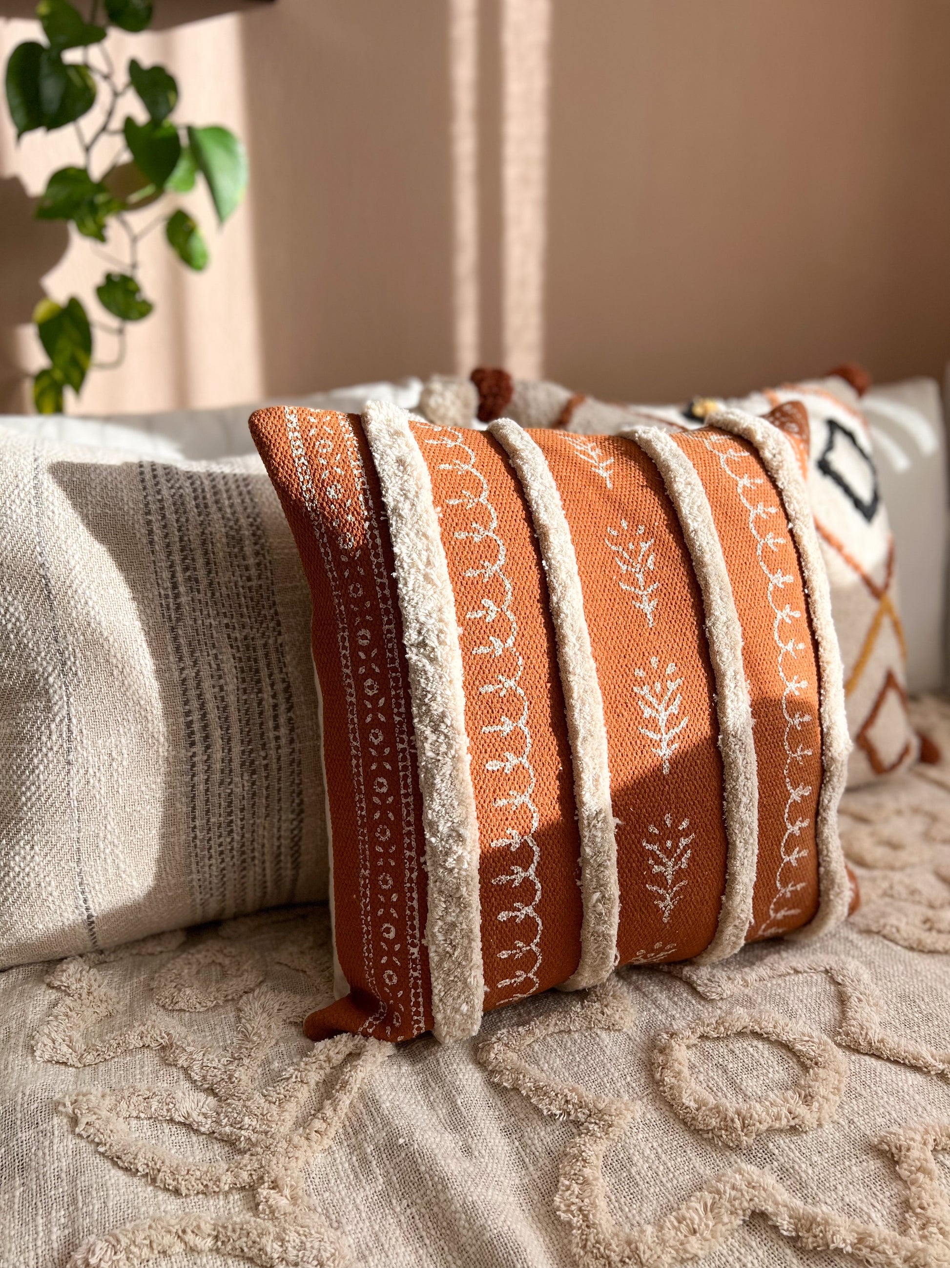 Lifestyle shot of the Nidhi tufted ochre cushion placed on a cozy bed alongside gray patterned decorative pillows in warm sunlight.