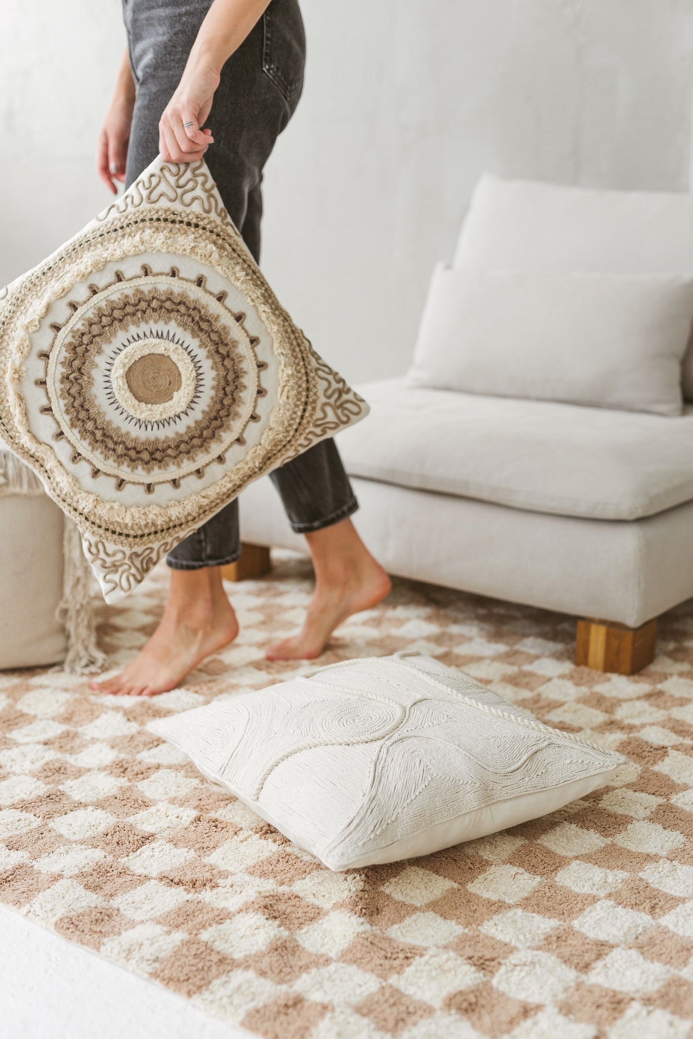 Lifestyle shot of a person holding the Saira artisanal cushion in front of a modern off-white armchair and checkered rug.