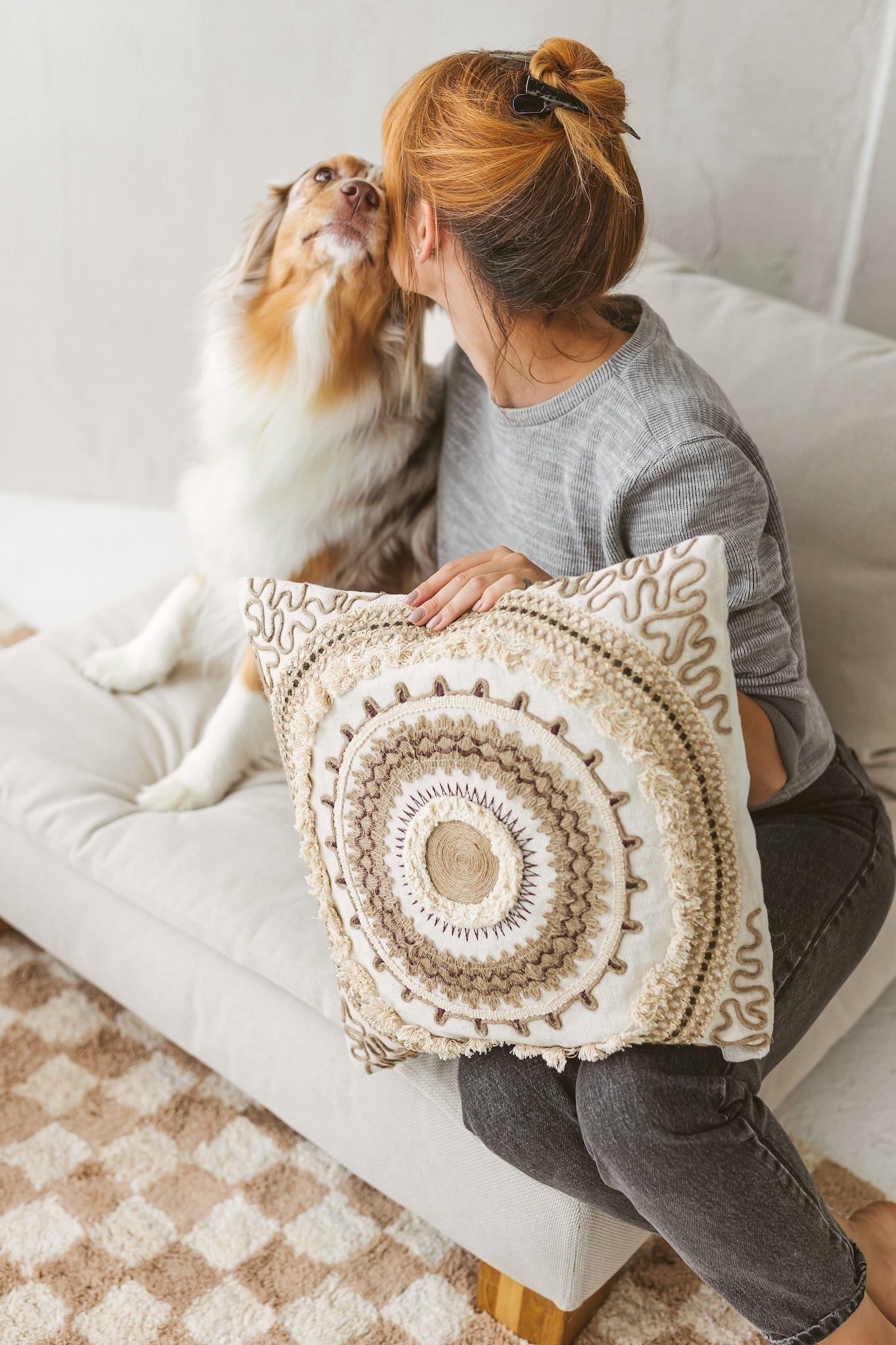 A woman sitting on a beige sofa with her dog, holding the highly textured Saira bohemian cushion with brown embroidery and tassels.