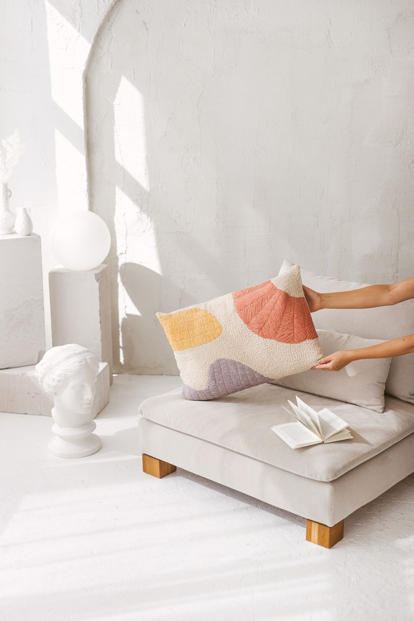 Lifestyle shot of the artisanal abstract cushion in a warm minimalist living room, styled next to a classical marble bust and ceramic vases in bright sunlight.